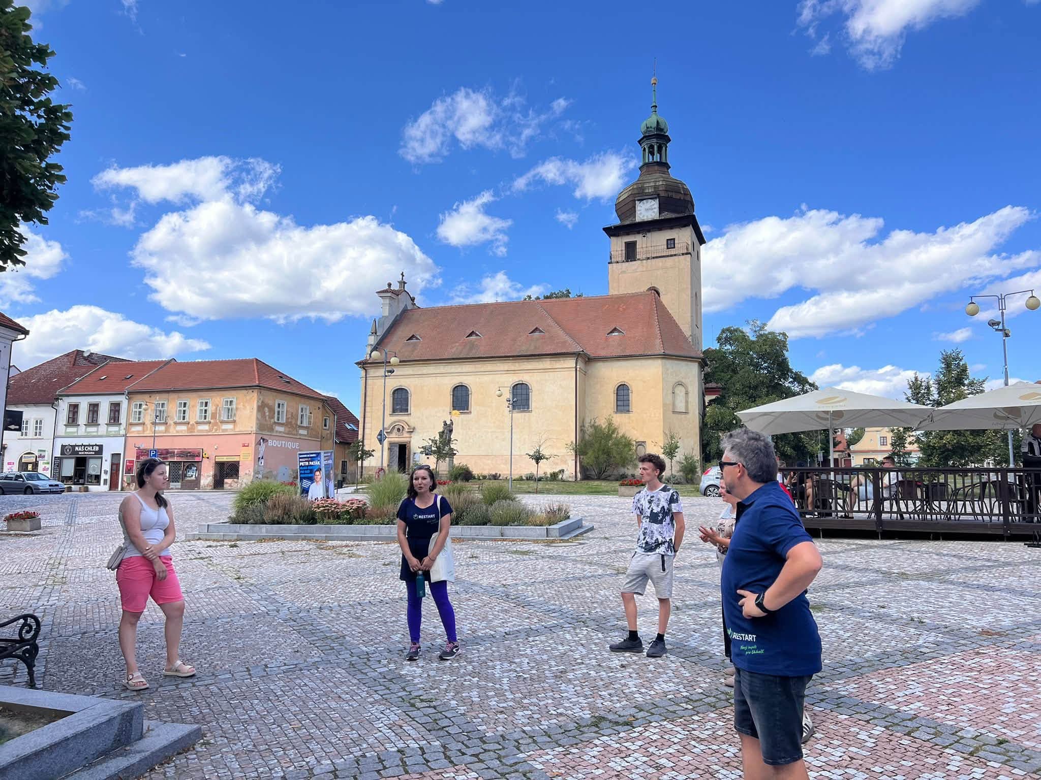 Účastníci komentované procházky stojí na Václavském náměstí v Unhošti před kostelem